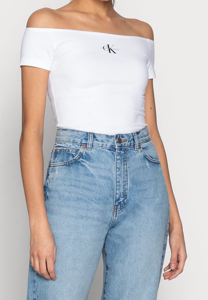Žena ve bílém off-shoulder topu od Calvina Kleina a v pase vysokých světle modrých džínech na jednobarevném pozadí.