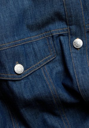 Blauwe denim stof met zichtbare stiksels, twee voorzakken beveiligd met zilveren metalen drukknoopjes, een gladde textuur en klassieke ontwerpelementen.