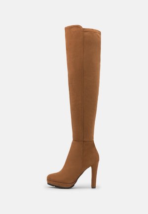 Botte haute en daim marron jusqu'au genou avec un bout arrondi, une semelle plateforme et un talon aiguille haut sur un fond clair uni.