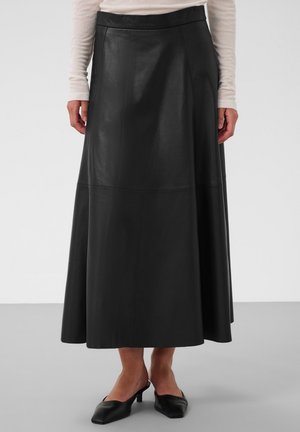 Femme portant une longue jupe en cuir noire, un haut beige à manches longues et des chaussures noires à talons bas, debout devant un fond uni.