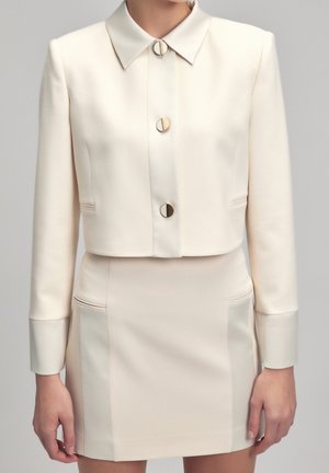 Afgekorte crèmekleurige blazer met gouden ronde knopen, voorzien van een puntkraag en zijzakken. Gecombineerd met een bijpassende aansluitende rok.