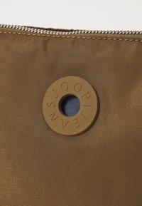 Detalj av en brun tygväska med blixtlås, prydd med en rund tan-färgad logotyp och en liten cirkulär blå öppning. Texturerad yta synlig.