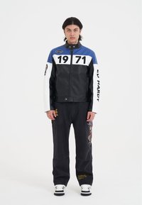 Svart läderjacka med blå och vita accenter, prydd med stora siffror "1971" på bröstet. Kombineras med svarta jeans och sneakers.