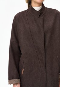 Kobieta ubrana w ciemnobrązowy wełniany płaszcz z wysokim kołnierzem, ukrytym zapięciem z przodu oraz beżowymi podwiniętymi mankietami, noszony na białym prążkowanym swetrze.