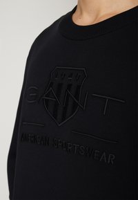 Fekete pulóver, textúrált pajzs emblémával, rajta "1949" és "GAINT AMERICAN SPORTSWEAR" sötét hímzéssel, bordás nyakkal.