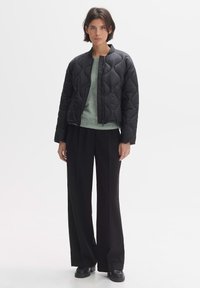 Gequilted zwarte bomberjack met een ritssluiting, gecombineerd met een lichtgroene trui, wijdvallende zwarte broek en zware zwarte schoenen.