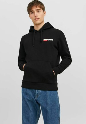 Sort hoodie lavet af bomuld med en frontlomme. Har et lille logo med røde og hvide accenter. Båret sammen med blå jeans.