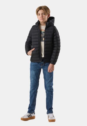 Junge trägt schwarze Steppjacke mit Kapuze, ein T-Shirt mit Grafik, blaue Jeans und weiße Turnschuhe mit Gummisohlen, steht vor einem einfarbigen Hintergrund.