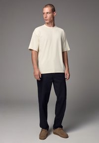 Beige cotton t-shirt, relaxed fit, short sleeves; paired with dark blue denim pants and brown suede loafers. Neutral background.
