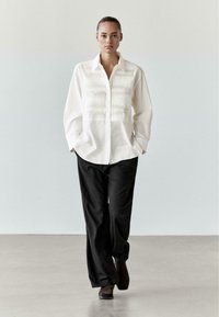 Bílá košile se zapínáním na knoflíky s texturovanými horizontálními záhyby, doplněná volnými černými kalhotami a hnědými kotníkovými botami. Hladká látka a uvolněný střih.