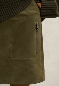 Fustă din piele întoarsă verde măsliniu, cu fermoar lateral și detalii de cusătură. Textura este moale, cu un finisaj neted și un design cu croială dreptă.