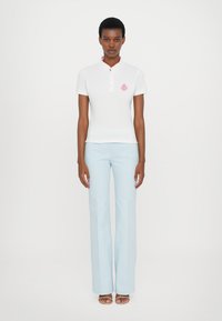 Hvit polo skjorte med rosa krage og logo, sammen med lyseblå vide bukser. Modellen står rett opp og ned, iført sandaler.