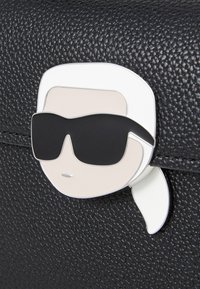 Borsa a tracolla in pelle nera texturizzata con un fermaglio in metallo che presenta un volto stilizzato con capelli bianchi e occhiali neri.