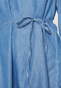 Vestido de denim azul claro com um cinto de laço na cintura. Possui uma textura suave e um tecido fluido, com um design plissado na parte da frente.