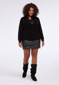 Cardigan nero lavorato a maglia con scollo a V e chiusura con bottoni, indossato sopra una camicia nera in satin e una minigonna in pelle nera, abbinato a stivali neri informali.