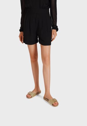 Shorts neri sartoriali con pinces e vita alta, abbinati a sandali dorati con fibbia decorativa e cinturini testurizzati.