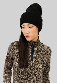 Bonnet noir en tricot côtelé avec un design slouchy. Porté avec un haut en polaire à imprimé léopard avec un col zippé noir.