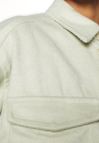 Camicia di flanella verde chiaro, strutturata, con colletto e una singola tasca sul petto. Il tessuto sembra morbido con una finitura leggermente spazzolata.