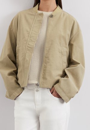 Persona con una chaqueta bomber beige con cremallera sobre un suéter blanco de punto y pantalones blancos de tiro alto, con las manos en los bolsillos.