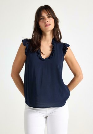 Mörkblå ärmlös topp med volangkrage och axeldetaljer, gjord av mjukt tyg, matchad med vita byxor. Enkel och elegant design.