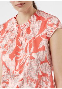 Koralowa bluzka z krótkimi rękawami, ozdobiona białym wzorem kwiatowym i subtelnymi fałdami w okolicy dekoltu. Gładka, lekka tekstura materiału.