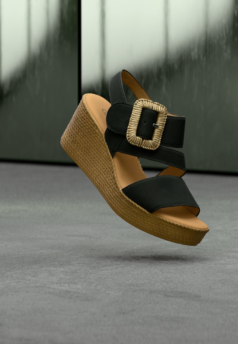 Sandale compensée noire avec semelle plateforme tissée marron et large bride à boucle tressée, flottant au-dessus d'une surface grise sur un fond sombre.