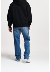 Sudadera negra con un corte relajado, combinada con vaqueros de mezclilla azul claro. Zapatillas blancas visibles en la parte inferior, que exhiben un diseño informal.