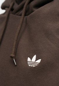 Camisola de capuz castanha com tecido texturado, capuz com cordão e um logo branco bordado em destaque com três riscas no peito.