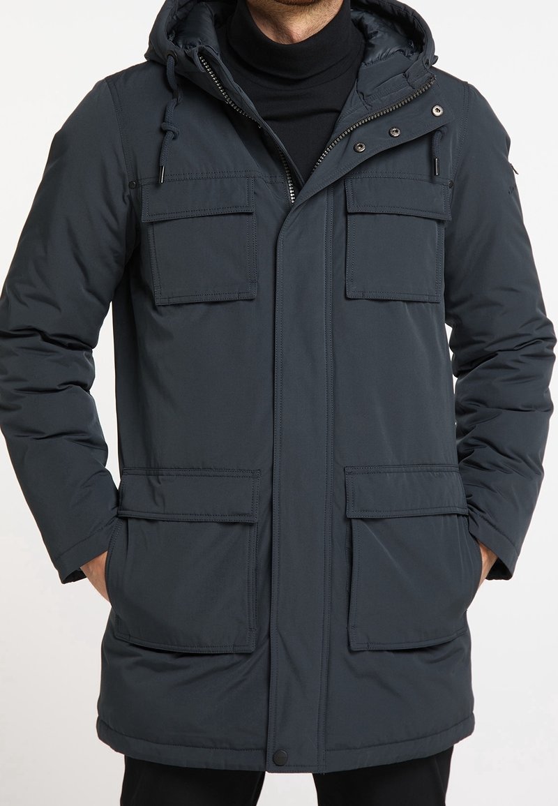 Homme portant une veste d'hiver à capuche bleu foncé avec quatre poches avant à rabat et les mains dans les poches inférieures.