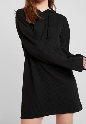 Kvinde iført en sort langærmet rillede hættetrøjekjole med snore, stående mod en ensartet lysegrå baggrund.