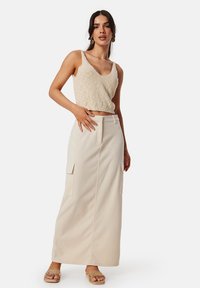 Beige gebreide crop top gecombineerd met een beige maxi rok met twee zijzakken en een rits aan de voorkant. Model staat op platte sandalen.