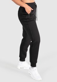 Person i svarta joggingsbyxor med dragkedjsidofickor och vita sneakers, stående mot en enfärgad ljusgrå bakgrund.