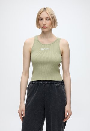 Femme avec coupe au carré blonde portant un débardeur côtelé vert clair et un pantalon noir ample, debout devant un fond blanc uni.