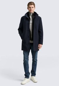 Temno modri plašč iz volnene mešanice z visokim ovratnikom, spredaj na gumbe in z dvema stranskima žepoma, oblečen preko belega pletenega puloverja, kombiniran s temnimi kavbojkami in supergami.