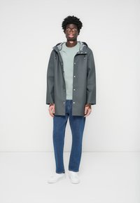 Grå vanntett regnjakke med hette, sølvfargede detaljer, brukt over en lys grønn skjorte, sammen med blå jeans og hvite joggesko.