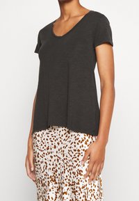 Eine Frau trägt ein kurzärmliges, dunkelgraues V-Ausschnitt-Oberteil, kombiniert mit einem weißen Rock, der braune Leopardenflecken aufweist.