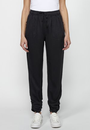 Pantaloni neri con elastico in vita, tasche laterali, orli affusolati e una texture liscia, abbinati a sneakers bianche.