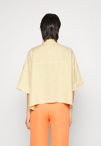 Person med kort sort hår iført en løs beige linnedskjorte og lyseorange bukser, der står foran en ensfarvet lys baggrund.