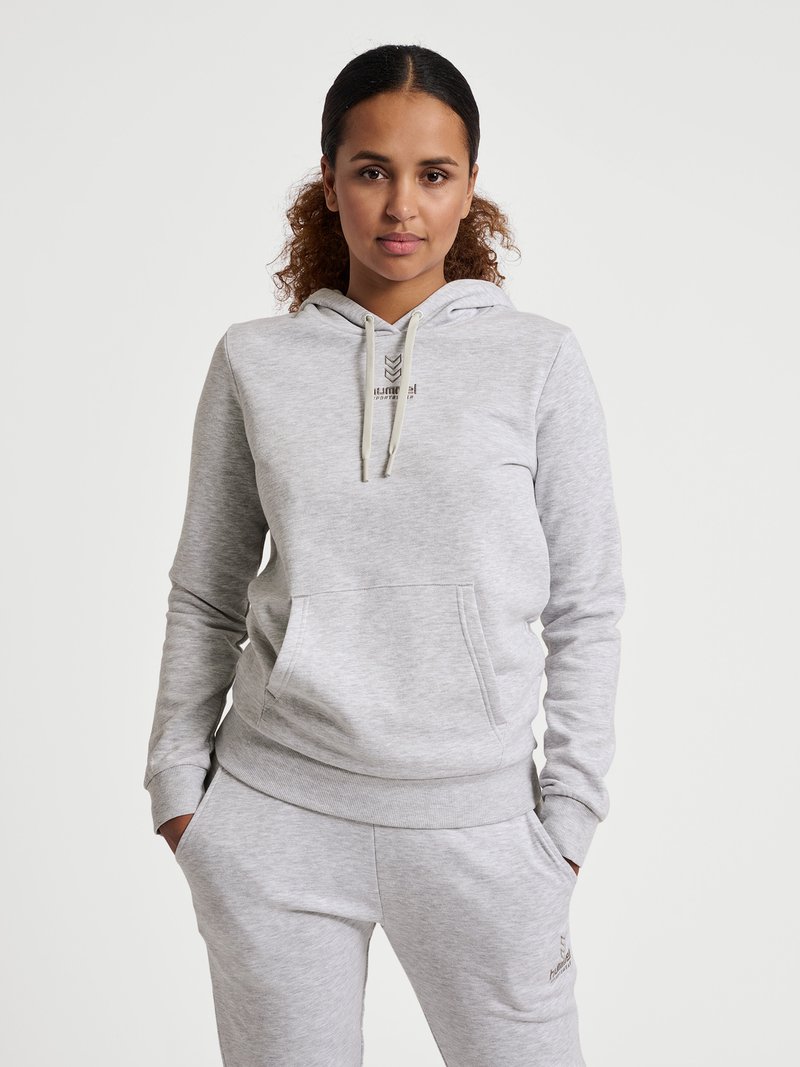 Frau mit lockigem Haar in einem hellgrauen Kapuzenpullover und passenden Jogginghosen steht mit den Händen in den Taschen vor einem schlichten Hintergrund.