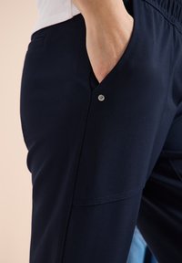 Personne portant un pantalon bleu marine foncé avec la main dans la poche avant, montrant la texture du tissu et un petit rivet en métal près du bord de la poche.