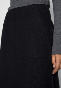 Jupe en laine noire avec une surface texturée, dotée de poches latérales et d'un design droit. Accompagnée d'un haut à manches longues gris.
