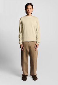 Felpa beige a maniche lunghe con collo rotondo, abbinata a pantaloni khaki larghi e scarpe slip-on marroni. Design minimalista con dettagli sottili.