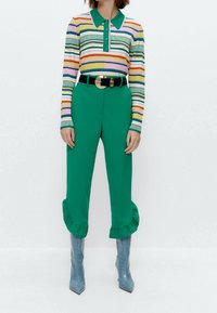 Gestreepte longsleeve polo in groene, roze en gele tinten, gecombineerd met groene textuurbroek met gerimpelde zoom en lichtblauwe puntige laarzen.