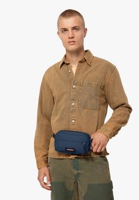 Marsupio blu navy con chiusura a zip e un logo, indossato sopra una maglietta beige con tasca. Materiale in cotone, design casual.