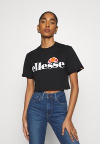 Schwarzes, kurzes T-Shirt aus Baumwolle mit einem großen weißen Logo und roten und orangefarbenen Akzenten. Getragen mit blauen hochgeschnittenen Jeans.