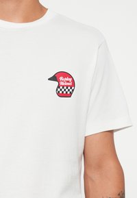 White t-shirt featuring a small red and black graphic of a helmet with checkered accents, displaying the text "Replay Helmet."