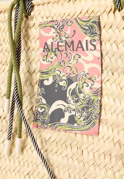 Surface en paille tissée avec une étiquette en tissu rectangulaire colorée présentant des motifs tourbillonnants verts, roses et noirs et le texte "ALEMAIS", et trois cordons.