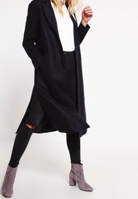 Langer, dunkelblauer Mantel mit Reverskragen, seitlichen Schlitzen und Knopfverschluss, getragen über einem weißen Oberteil und schwarzen Jeans. Graue Wildleder-Stiefeletten.