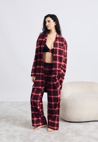 Σετ πιτζάμες από φλαανέλ με κόκκινο και μαύρο καρό σχέδιο, που αποτελείται από άνετο πουκάμισο και παντελόνι. Μαλακή υφή, μαύρο ρυθμιζόμενο αθλητικό σουτιέν και γυμνά πόδια.