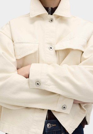 Personne les bras croisés portant une veste en denim crème par-dessus un haut foncé et un jean bleu avec une fermeture éclair et un bouton visibles.